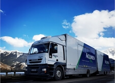 side view of an Acumen covered car transport lorry driving along a road with a picturesque mountain in the background.